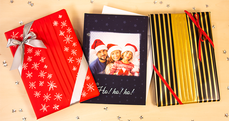 2 photobooks wrapped in a fan-style gift box, a photobook with a navy blue cover and a photo of a smiling family wearing Santa Claus hats in the middle. Silver stars all around them.