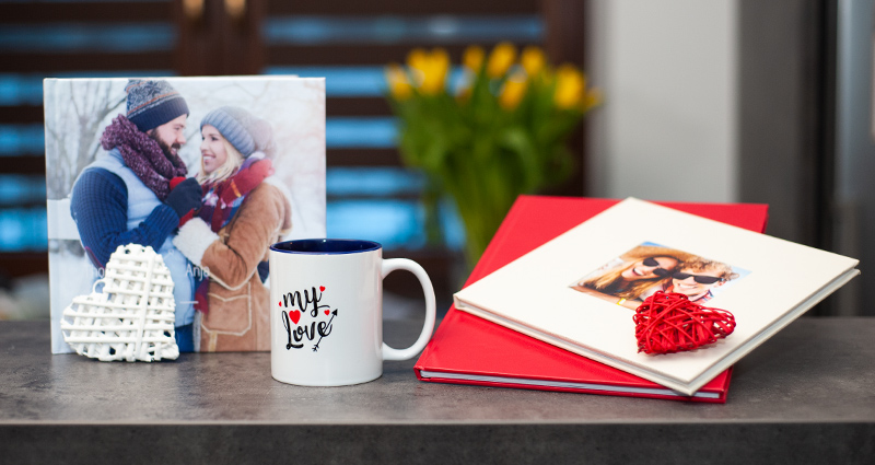 3 photo book types (2 exclusive photo books and 1 classic photo book) on a dark table, a photo mug and a decorative white heart next to them; yellow tulips in a bucket in the background.