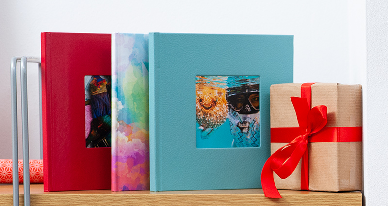 Drei quadratische Fotobücher auf einem Holzregal (2x Fotobuch Exklusiv und 1x Fotobuch Classic), daneben ein Geschenk mit rotem Band.