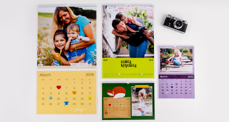 4 Fotokalender in verschiedenen Formaten auf weißem Hintergrund, daneben eine Kamera.