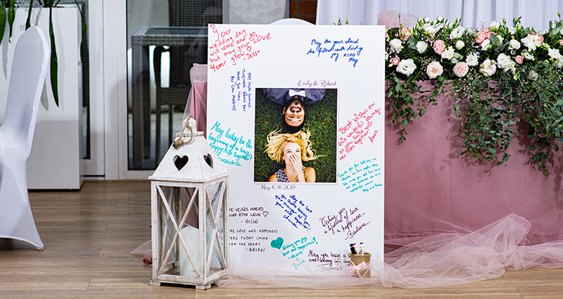 Een groot canvas met een foto van een verliefd paar in het midden; rondom - wensen van bruiloftsgasten geschreven met kleurrijke markeerstiften; ernaast een witte lantaarn en op de achtergrond - een tafel versierd met poederroze tule en bloemen.