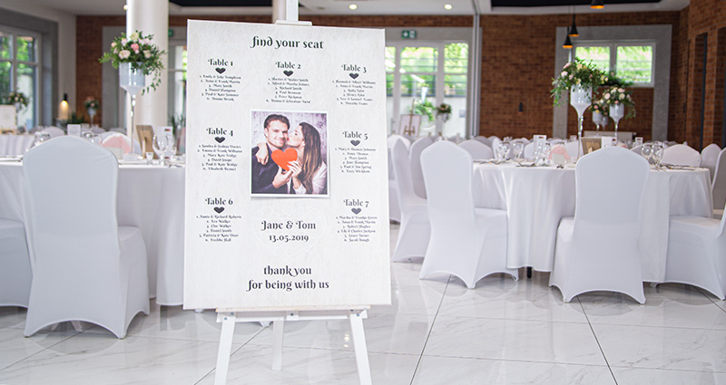 A big photo canvas with a photo of the bride and groom on an easel in the middle of a ballroom. A list of wedding guests’ surnames near specific table numbers around the photo. Tables and chairs decorated in white in the background.