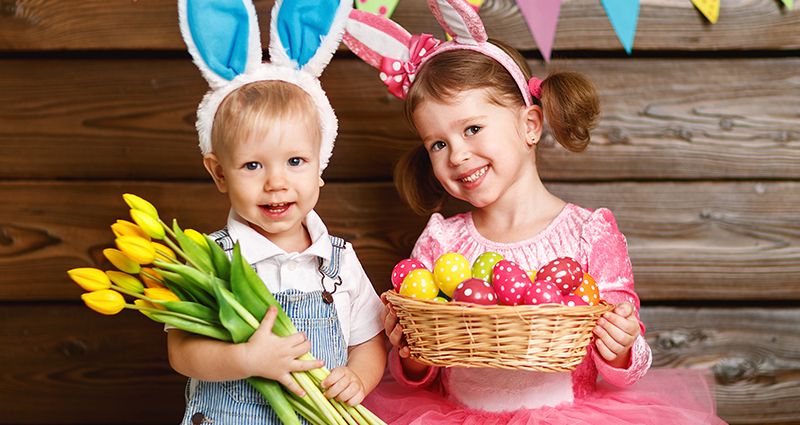 Een jongen en een meisje dragen hoofdbanden die op konijnenoren lijken. De jongen houdt een boeket tulpen en het meisje een mand met paaseieren. Op de achtergrond is er een houten muur en een gekleurde slinger.