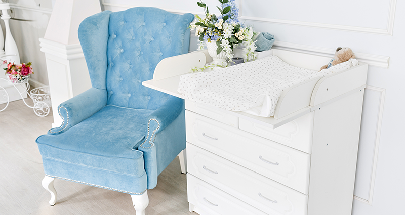 A changing table with a protective mat on the chest of drawers, a blue armchair next to it