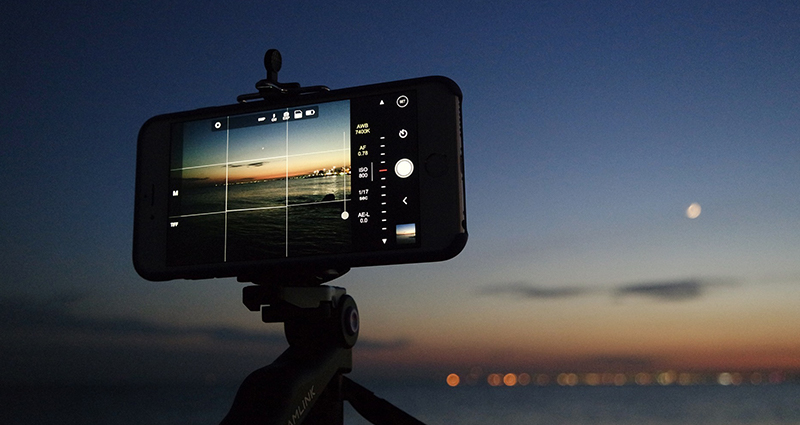 Zoom in op een smartphone die wordt bevestigd aan een statief. Op het scherm het strand en zee bij nacht. 