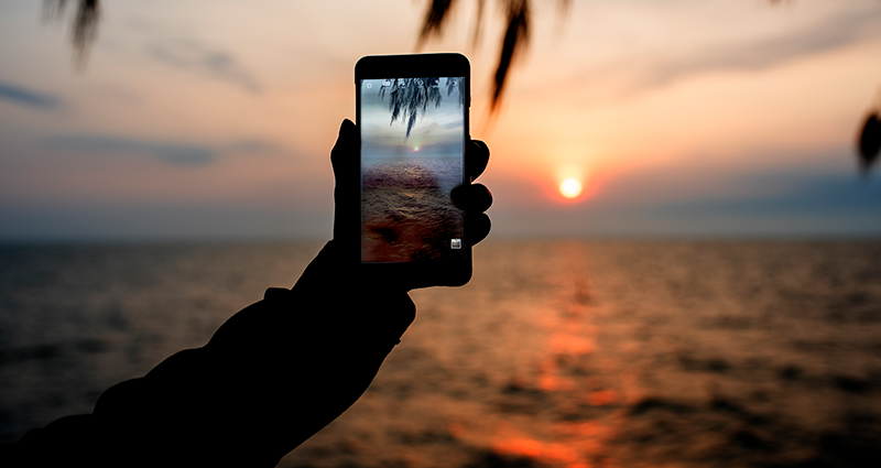 A close up on a man’s hand holding a smartphone and taking a photo of a sunset by the sea.