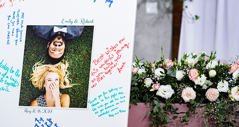 Detail velkého plátna s fotografií zamilovaného páru uprostřed, kolem přání od svatebních hostů psaná barevnými fixy. Na pozadí, na stole pudrově-růžové barvy, kytice květů v pastelových barvách.