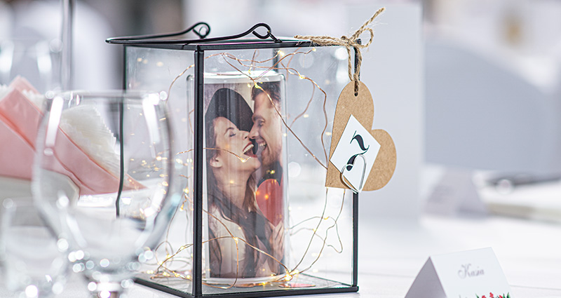 Nachaufnahme einer Glaslaterne, die auf dem Hochzeitstisch mit einem aufgerollten Foto des verliebten Paares in der Mitte und mit den LED-Lampen steht. An der Laterne angebrachte Nummer 7.