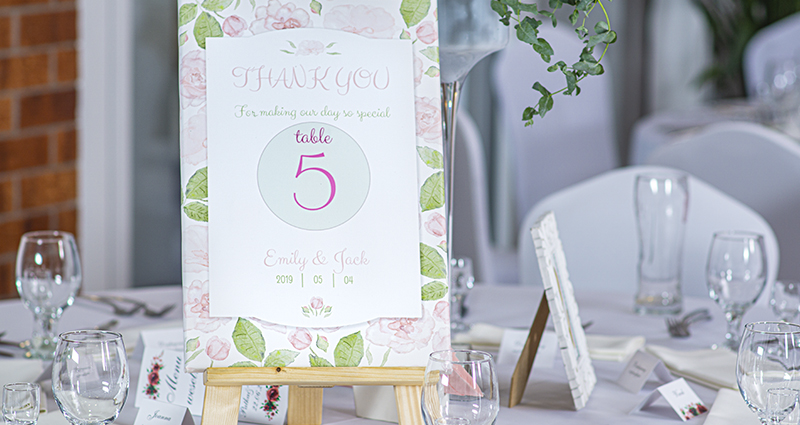 Nahaufnahme einer pastellfarbene Fotoleinwand mit der Tischnummer und den Dankessprüchen für die Gäste auf einem weiß verzierten Hochzeitstisch.