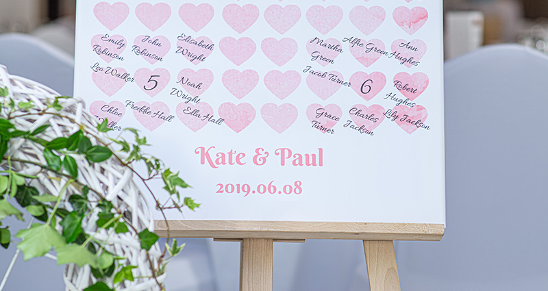 Close-up op een tafelindeling welkomstbord (Aquarel roze-sjabloon met kleine roze hartjes); onderaan het sjabloon de namen van het bruidspaar en de datum van de bruiloft. 