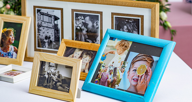 A close-up on colourful frames with photos of the groom, the bride and their families from different stages of life, placed on a table decorated with flowers