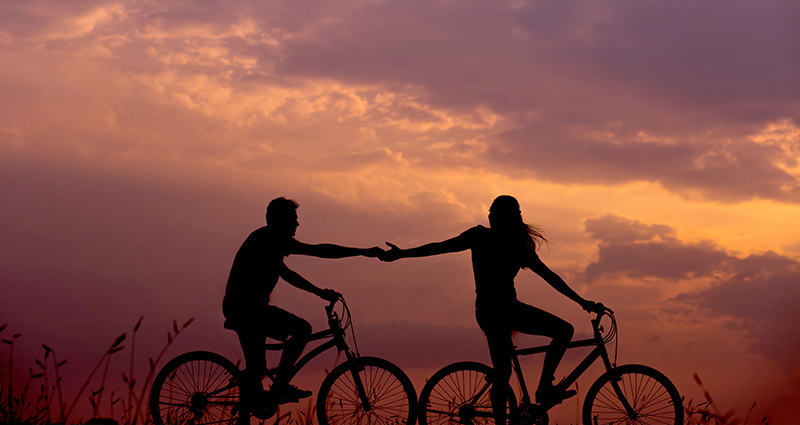 A couple riding on bikes and holding their hands; sunset in pink and orange shades in the background.