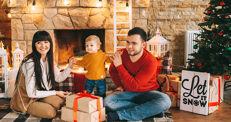 A couple with a small kid sitting on a blanket in front of the fireplace decorated with lights. A Christmas tree and gifts next to them.
