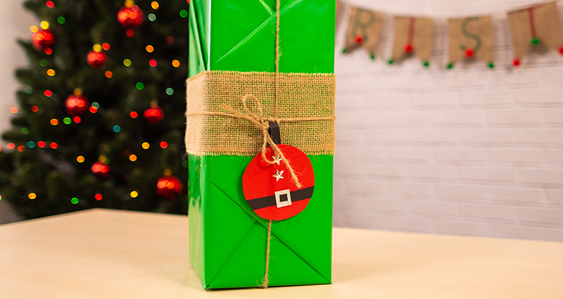 Un cadeau emballé en papier couleur verte en style kimono avec une étiquette en forme de Père Noël. au fond le texte Merry Christmas et un arbre de  Noël.