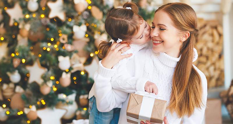 Mergaitė bučiuoja mamą, turinčią dovaną į skruostą, fone yra Kalėdų eglutė.