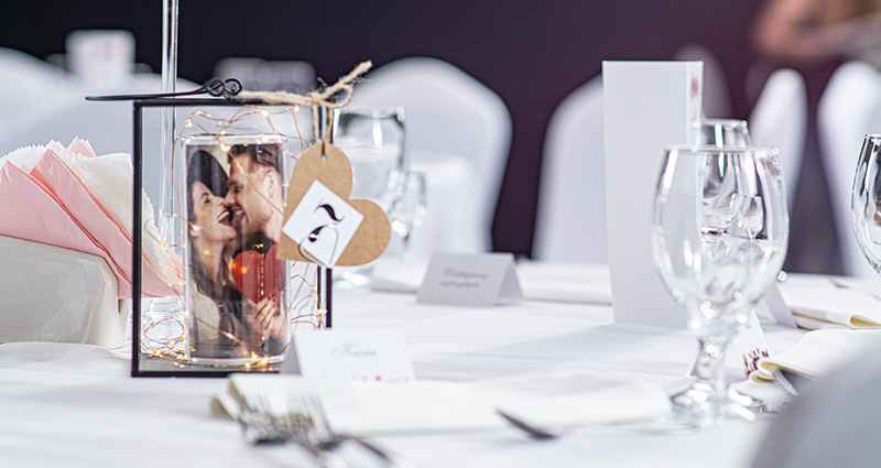 A glass lantern with rolled photo of a couple in love inside and led lights. Number 7 attached to the lantern. The lantern set on a decorated white wedding table. Tableware around.