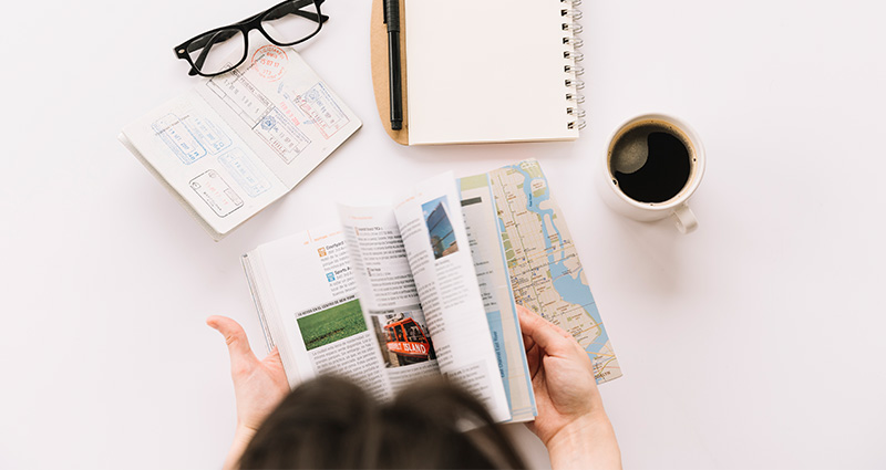 Flatlay-Foto: Frau, die in einem Reiseführer blättert. Auf dem Schreibtisch befindet sich ein Pass, ein Notizbuch und eine Tasse Kaffee. 