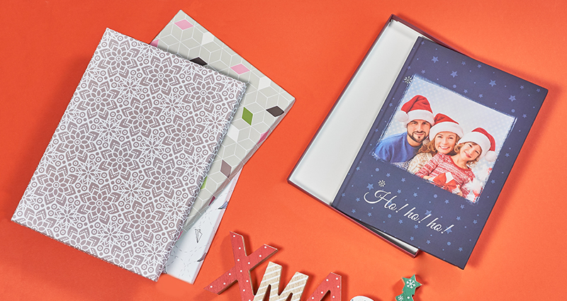 A photo book with a navy blue cover showing a smiling family wearing Santa Claus hats; 3 gift boxes for photo books placed in a pile and a wooden letters XMAS below. All products are lying on the red background.