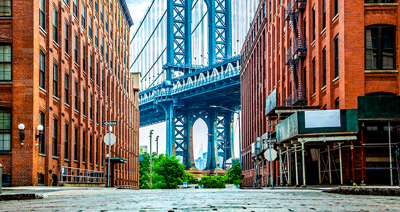 Ein Foto der Manhattan Bridge, das zwischen zwei Backsteingebäuden und aus der Forschperspektive aufgenommen wurde. 
