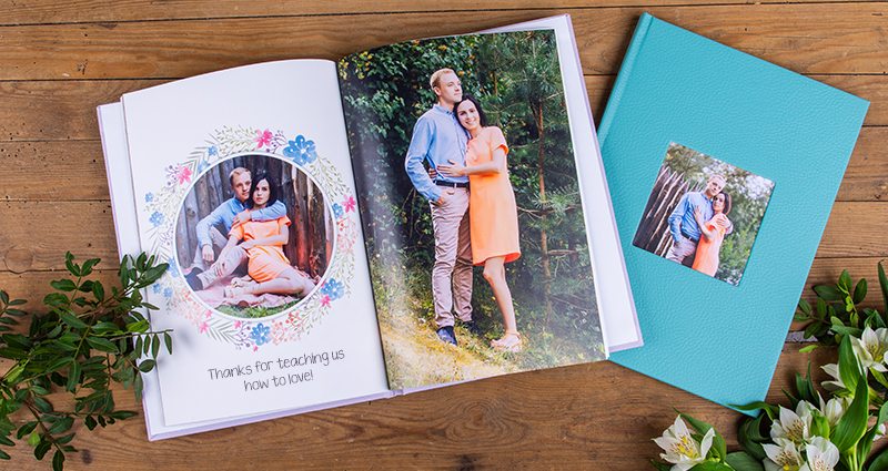 A picture of 2 photo books on a dark wooden background, flowers and decorative plants on both sides. One of the books is open, on its pages pictures of an engaged couple and a caption: 'Thanks for teaching us how to love!'. Closed photo book with a turquoise eco leather cover and a photo of the same couple in a photo slot. 