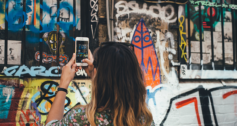 A picture of a woman taking a photo of a graffiti, with her smartphone, on a town house’s wall.