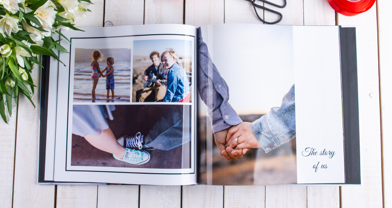 A picture of an open square photo book with pictures of a couple starting from their childhood till present. A photo book on a bright wooden background, red ribbon and scissors above that. White flowers bouquet next to it.