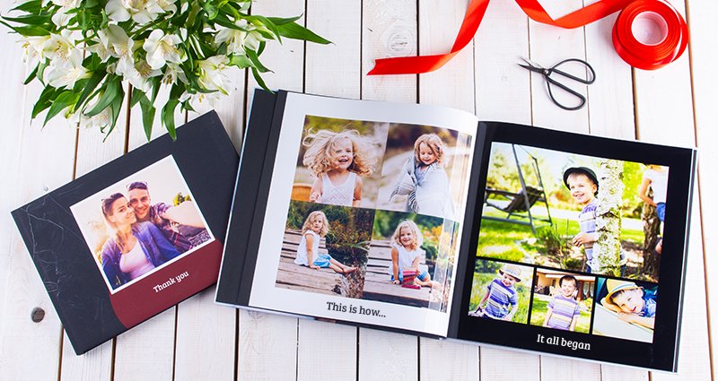 A picture of two photo books – one of the albums with the pictures of a couple and 'Thank you' caption is closed, an open photo book next to it (on the left side page photos from girl's childhood and boy's childhood on the right). Red ribbon and scissors above the open album. White flowers bouquet in the upper left corner. 