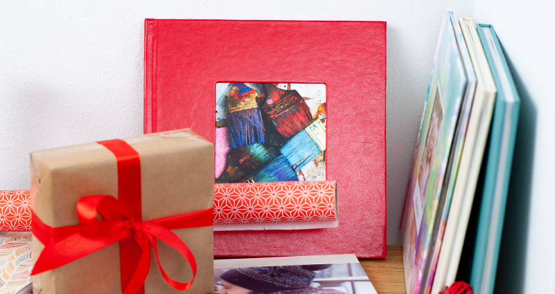 Priartinimas ant esančios lentynos su įvairiomis fotoknygų rūšimis ir formatais, šalia dovana, supakuota į pilkos spalvos popierių ir surišta raudona juosta.