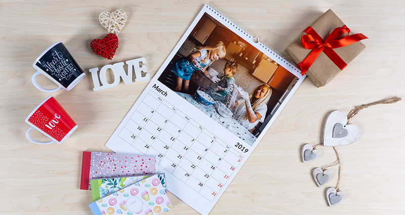 A4 fotokalendorius su žaidžiančios šeimos virtuvėje nuotrauka, šalia 3 sharebookai, du puodeliai ir dovana su raudona juosta ir dekoratyvine širdimi.