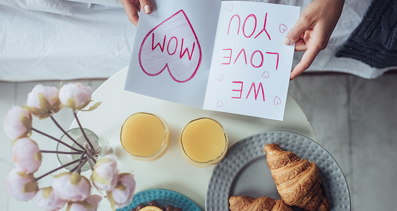 Aerial view – a close up on a card with the “WE LOVE YOU MOM” caption. Nearby a bouquet of light flowers, two glasses of orange juice and two croissants.