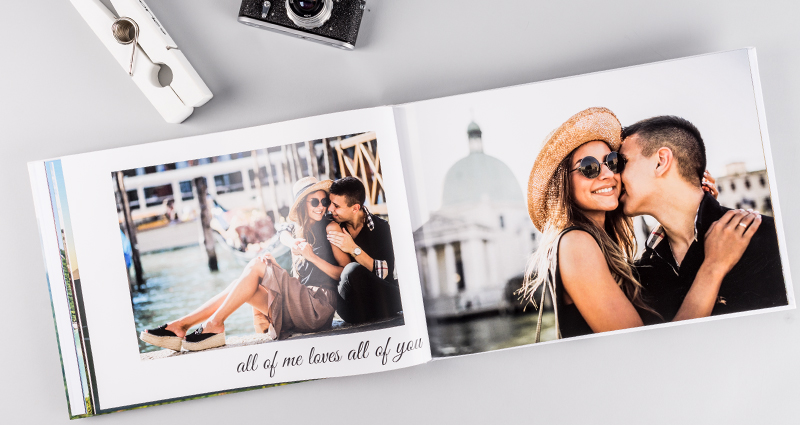 An open photo book A3 landscape, with pictures of a couple in love, lying next to a big paper clip and a camera