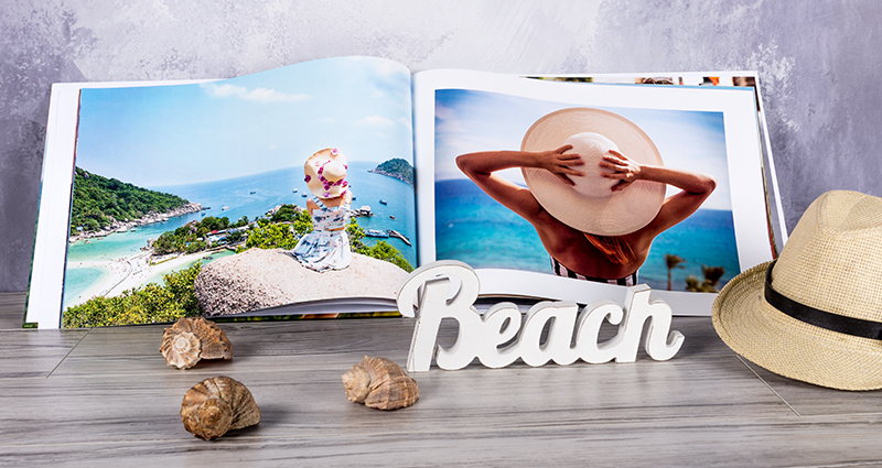 Fotolibro A3 aperto con la foto di una donna al mare. Accanto delle conchiglie, un cappello e un’incisione in legno con la scritta "Beach"