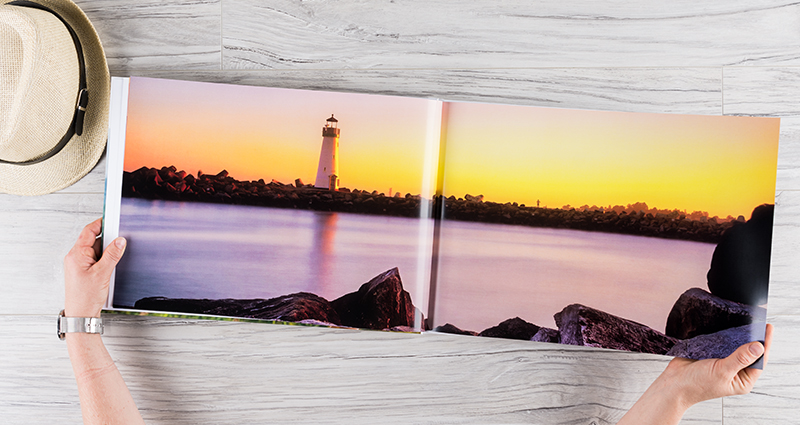 Fotolibro A3 abierto sobre un escritorio con una foto horizontal de la orilla del mar impresa en dos páginas, un faro en el fondo. Un sombrero al lado del libro.