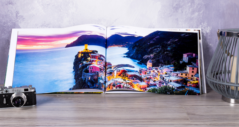 Un livre photo A3 ouvert  posé sur une table. Sur la photo du livre la vue de la mer. A côté du livre un lampadaire  et un appareil photo.