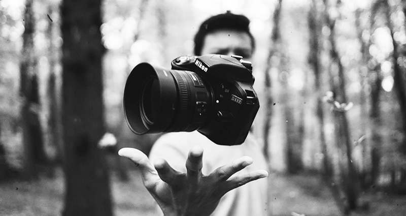 Black and white photograph showing a man in the woods throwing a camera up