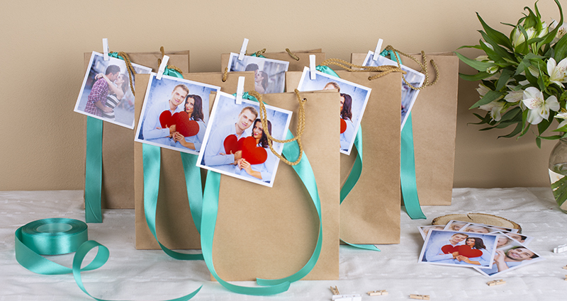 Paper bags decorated with turquoise ribbon and insta photos of a couple attached with a tiny wooden clips. Prints, scissors, spool of ribbon and a white flowers bouquet in a vase. A composition on a white tablecloth. 
