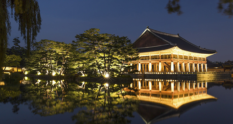 Japoniško stiliaus pastatas ir ežere atspindintys medžiai, naktį fotografuota nuotrauka.