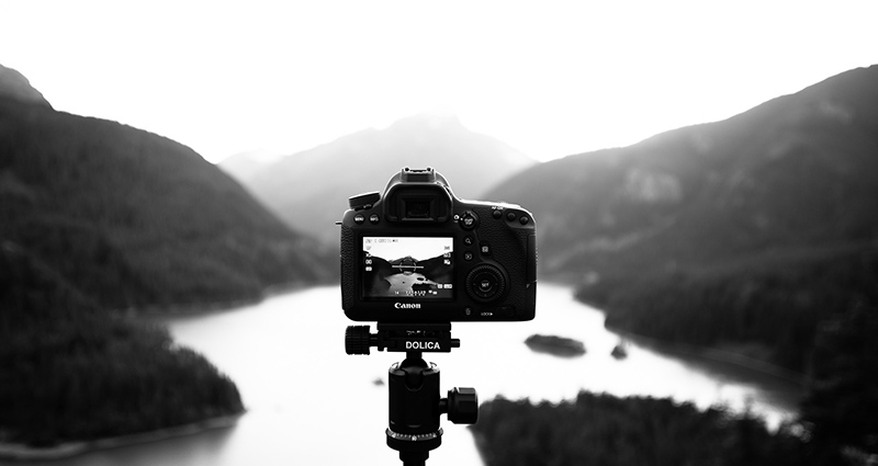 Fotoaparatas ant trikojo, fone ežeras ir kalnai - juodai balta nuotrauka.