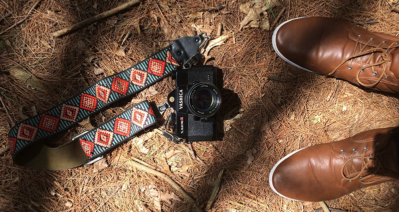 Una cámara fotográfica con una correa hecha a mano dispuesta en la tierra, un enfoque a los zapatos marrones de hombre; foto tomada a vista de pájaro.