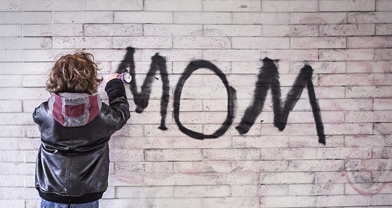 Niño pintando la palabra "MOM" con spray en la pared. 
