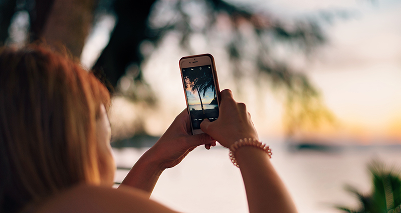 Detailní záběr na ženu, která fotografuje se smartphonem palmy na pláži, v pozadí moře a slunce.