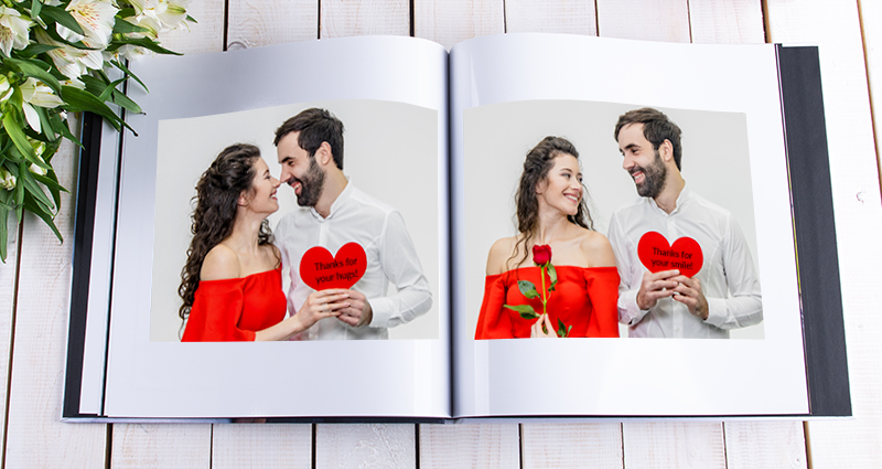 Nahaufnahme eines aufgeschlagenen Fotobuches (Starbooks) mit Fotos eines verliebten Paares, das die Papierherzen mit Aufschriften hält. Links ein Strauß weißer Blumen, in der oberen rechten Ecke ein verpacktes buntes Geschenk.