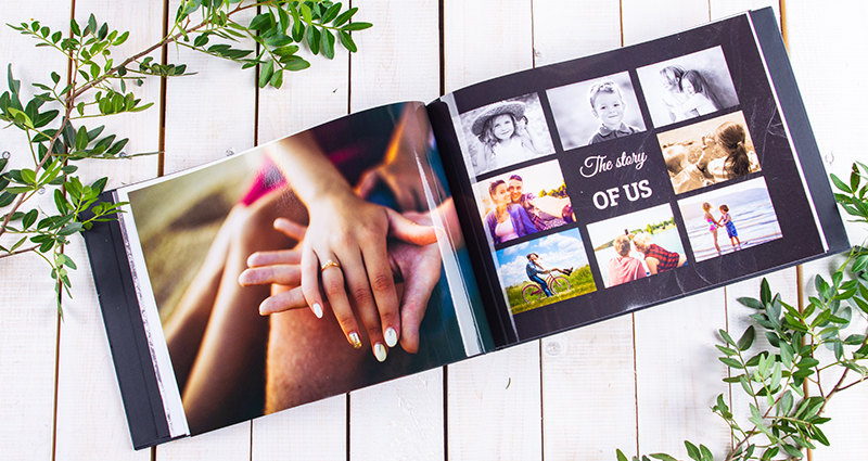 Detailní záběr na otevřenou fotoknihu - na levé straně fotografie rukou snoubenců, na pravé koláž z fotografií. Fotokniha leží na světlém dřevěném pozadí se zelenými větvemi.