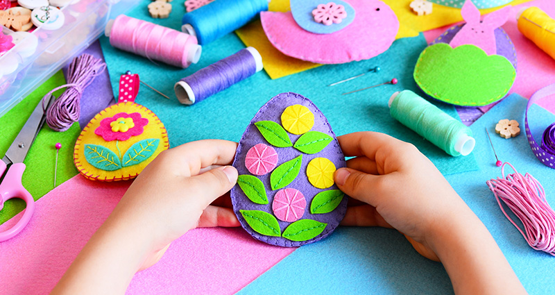 Close-up on a child's hands holding a colourful felt Easter egg. Threads and other felt Easer decorations in the background.