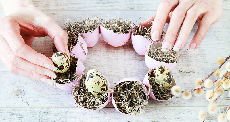 Zoom sobre la mano de una mujer que está haciendo una decoración de Pascua utilizando unas cáscaras de huevos, un musgo y un amento.
