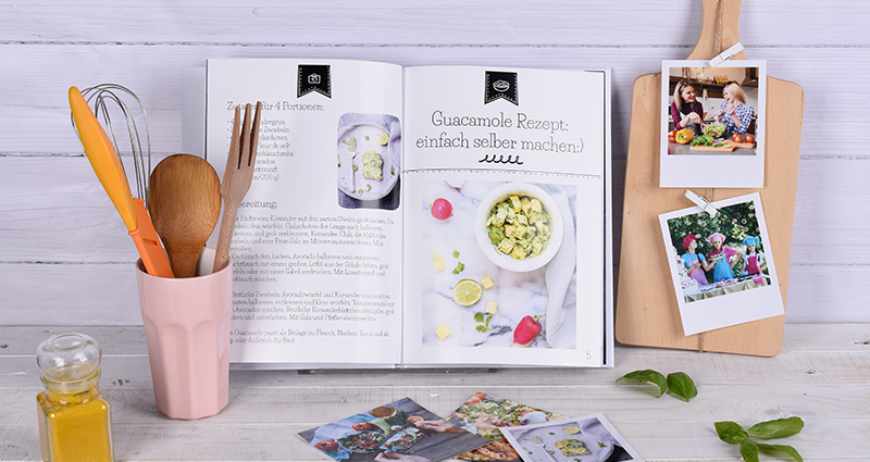 Cookbook lying next to retro prints on a chopping board; cooking utensils in a mug next to photos