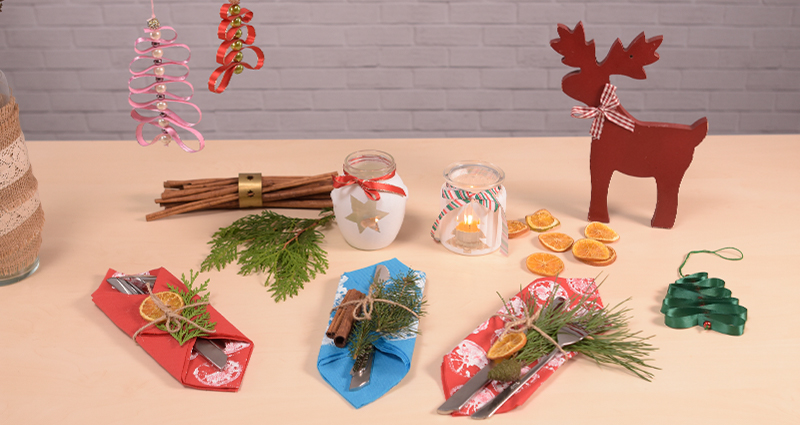 DIY-Weihnachtsdekorationen: dekorative Besteckservietten und Laternen aus Glas auf einem hellen Tisch neben Thuja-Zweigen, Zimtstangen und Orangenscheiben. Daneben Zweige in einer Vase mit Leinenband dekoriert, und an ihnen Weihnachtsbäume aus Bändern