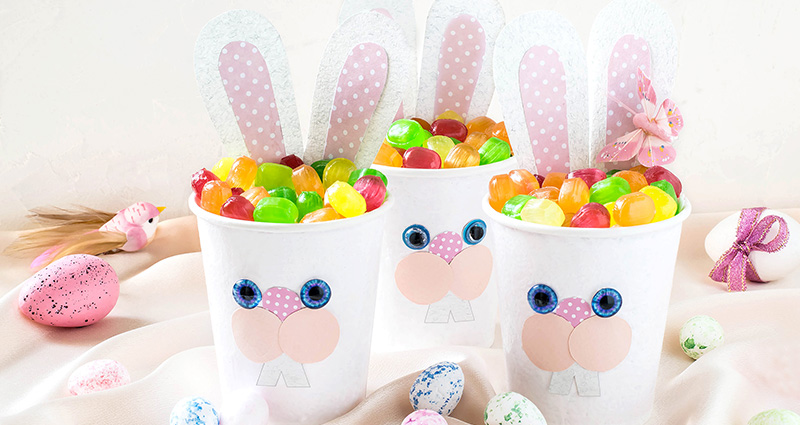 DIY zajačikovia z plastových pohárov plné sladkostí, okolo nich farebné veľkonočné vajíčka