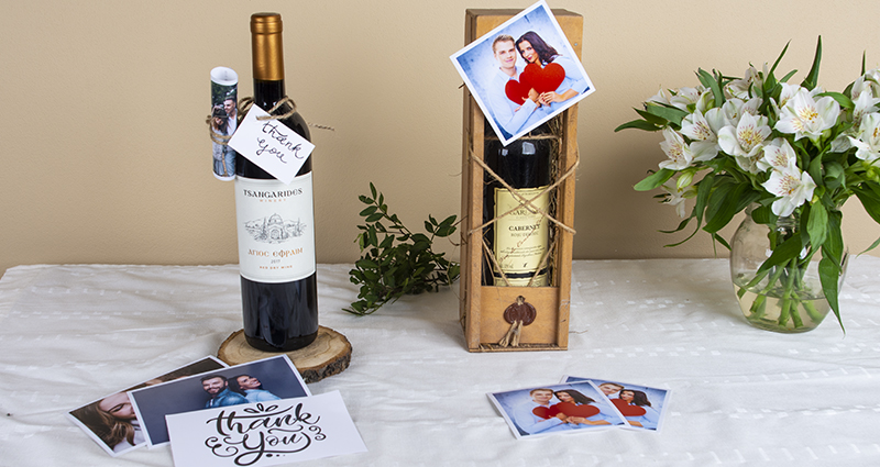Two wine bottles decorated with prints of a couple and a card saying 'Thank you', prints and insta photos placed next to them and also white flowers bouquet in a vase. A composition on a white tablecloth. 