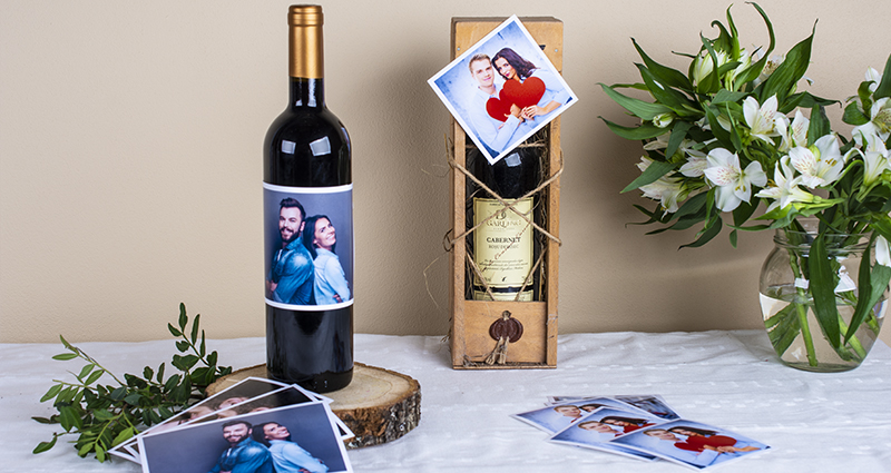Two wine bottles decorated with prints of a couple, prints, insta photos and whithe flowers bouquet in a vase placed next to them. A composition on a white tablecloth.